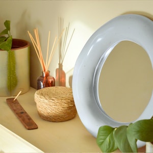 May include: A white, oval-shaped mirror with a light-colored frame is the focal point of this image. Other items include a woven basket, reed diffusers in amber bottles, and potted plants. The scene is set on a light-colored shelf.