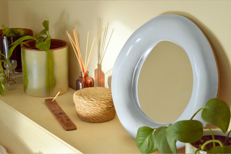 May include: A white, oval-shaped mirror with a light-colored frame is the focal point of this image. Other items include a woven basket, reed diffusers in amber bottles, and potted plants. The scene is set on a light-colored shelf.