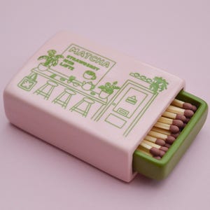 May include: A pink matchbox with a green interior, featuring a cafe-themed illustration and the text "Matcha Strawberry & Latte." The matchbox contains several matches with brown tips.