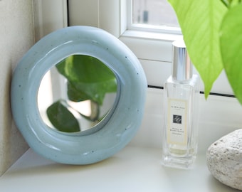 May include: A light blue, round ceramic mirror with a speckled finish. A clear perfume bottle with a silver cap and a small, smooth stone are next to the mirror. Green leaves are visible in the background.