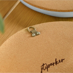 May include: Close-up of the back of a round, light brown corkboard with a metal hanger attached. The corkboard has a textured surface and the handwritten signature "jtparker" in black ink. A second corkboard with a blue rim is visible in the background.