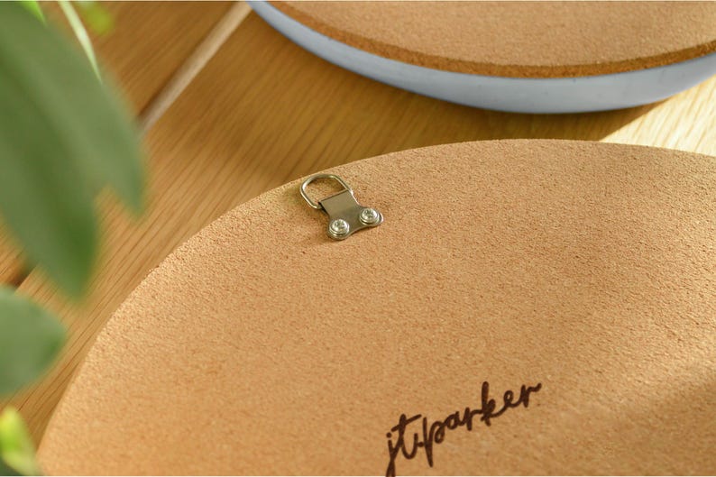 May include: Close-up of the back of a round, light brown corkboard with a metal hanger attached. The corkboard has a textured surface and the handwritten signature "jtparker" in black ink. A second corkboard with a blue rim is visible in the background.