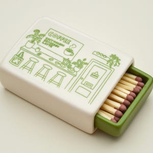 May include: A rectangular, white ceramic matchbox with a green interior. The lid features a green illustration of a coffee shop scene with the word "COFFEE". The matchbox is filled with wooden matches with red tips.