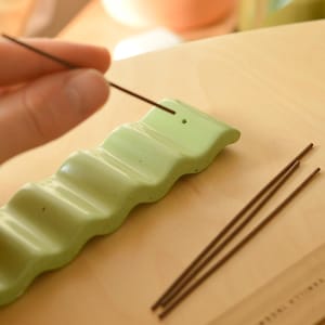 May include: A light green ceramic incense holder with a wavy design. The holder has multiple slots for incense sticks. A hand is holding a brown incense stick over the holder.