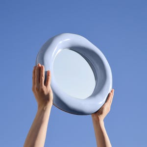 May include: A light blue, oval-shaped mirror is held up against a clear blue sky. The mirror has a thick, rounded frame. The hands holding the mirror are visible. The mirror's surface reflects the sky.