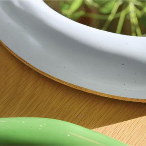 May include: Close-up of a white, round object with a light brown edge, possibly a plate or a decorative item. A green object is visible in the foreground. The objects are on a wooden surface.