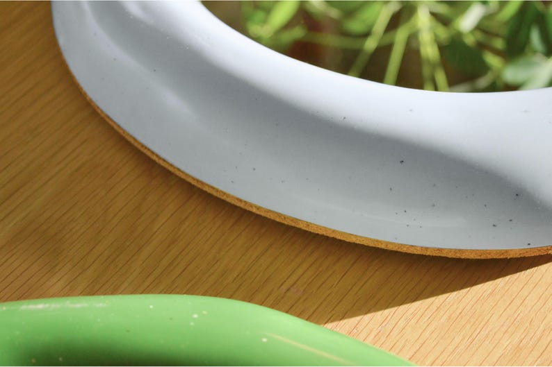 May include: Close-up of a white, round object with a light brown edge, possibly a plate or a decorative item. A green object is visible in the foreground. The objects are on a wooden surface.