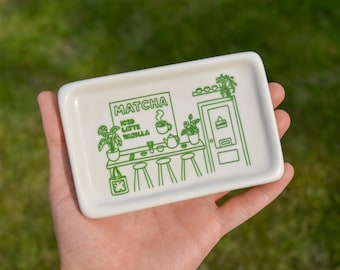 May include: A rectangular, white ceramic tray with a green line drawing of a matcha cafe scene. The design includes the word "Matcha", a counter with stools, plants, and a door with a "closed" sign.