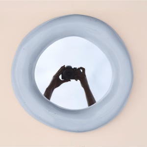 May include: A round, light blue mirror with an organic, uneven edge. The reflection in the mirror shows a person holding a camera. The mirror is mounted on a light peach-colored wall, creating a minimalist aesthetic.