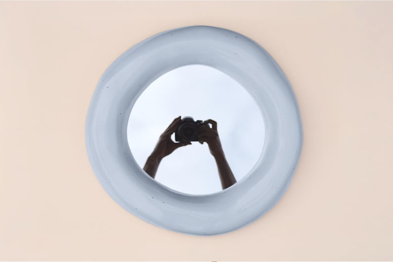 May include: A round, light blue mirror with an organic, uneven edge. The reflection in the mirror shows a person holding a camera. The mirror is mounted on a light peach-colored wall, creating a minimalist aesthetic.