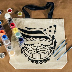 May include: A beige canvas tote bag with a black and white graphic of a smiling face with a mustache. Paint pots in various colors and paintbrushes are next to the bag. The tote bag has black handles.