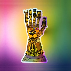 May include: A holographic sticker depicting a golden gauntlet with superhero characters on each finger. The gauntlet is highly detailed, with the text "MANO PODER USA" at the base. The background has a colourful gradient.
