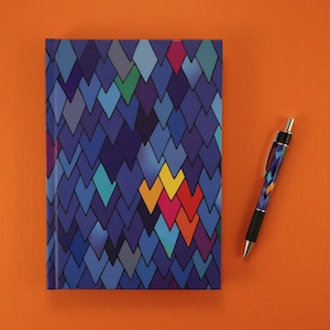 May include: A patterned notebook and pen set. The notebook features a blue diamond pattern with accents of teal, green, yellow, orange, and pink. The pen has a matching design. The set is displayed on an orange background.
