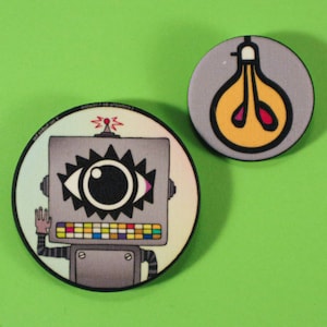 Puede incluir: Dos pines redondos y coloridos con diseños de dibujos animados sobre un fondo verde. Un pin presenta un robot con un ojo grande, el otro una forma de bombilla. El pin del robot tiene el texto "Conquer or Collide" en el borde.