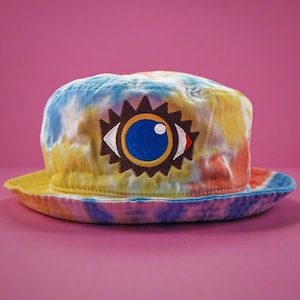 May include: A tie-dye bucket hat with a vibrant mix of yellow, blue, and orange hues. The hat features an embroidered eye design with a blue iris, brown outline, and a red accent. The brim is approximately 2.5 inches wide.