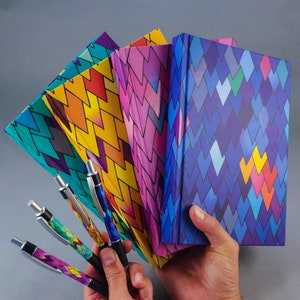 May include: A collection of notebooks and pens with matching colorful geometric patterns. The notebooks are fanned out, showcasing vibrant hues of blue, yellow, pink, and purple. The pens have black barrels and silver accents.