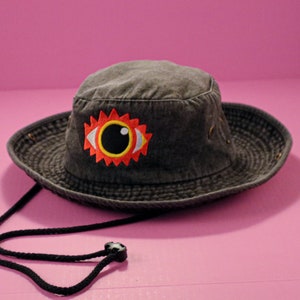 Puede incluir: Un sombrero de pescador gris oscuro con una correa de barbilla negra. El sombrero presenta un diseño de ojo bordado colorido con acentos rojos, amarillos y blancos. El sombrero está hecho de una tela texturizada y tiene ojales metálicos alrededor del borde.