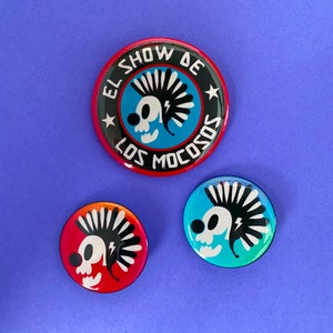 Puede incluir: Tres botones redondos con un diseño de calavera de dibujos animados. El botón más grande tiene el texto "EL SHOW DE LOS MOCOSOS" en blanco sobre un fondo negro con un borde rojo. Los otros dos botones tienen un diseño de calavera similar con un fondo degradado.