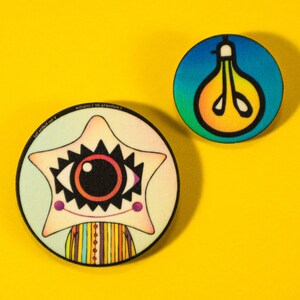 May include: Two colorful, round pinback buttons on a yellow background. One button features a star-shaped character with an eye and a striped body. The other button has a lightbulb design with a gradient of blue, green, and yellow.