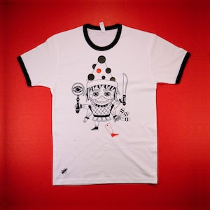 Puede incluir: Camiseta blanca con ribete negro y una ilustración caprichosa. El gráfico presenta un personaje de dibujos animados sosteniendo una lupa, un cuchillo y malabares. También se representa una bota roja. Una pequeña araña negra está en la parte inferior.