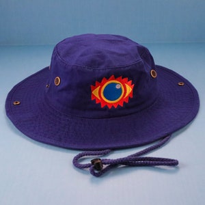Puede incluir: Un sombrero de pescador azul marino con ala ancha y correa para la barbilla. El sombrero presenta un diseño de ojo bordado colorido en la parte delantera. El diseño del ojo tiene un sol rojo y amarillo alrededor de un ojo azul y blanco.