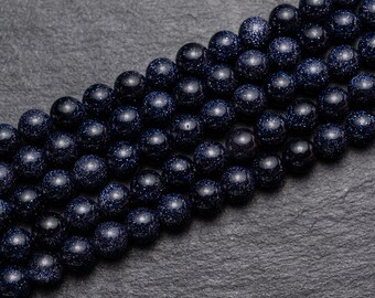 May include: Close-up of several strands of dark blue, round gemstone beads. The beads have a speckled, glittery appearance, with tiny flecks of lighter blue and silver. The beads are arranged in neat rows against a dark gray background.
