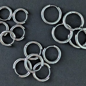 Pode incluir: Vários pares de brincos de argola prateados estão dispostos sobre uma superfície escura. Os brincos são circulares com um fecho articulado. Os brincos variam em tamanho e são feitos de um material metálico brilhante.