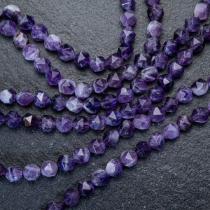 Può includere: Un filo di perle di pietra preziosa ametista, sfaccettate e lucidate, con un design a forma di stella. Le perle sono infilate su un filo trasparente e sono disposte in uno schema sciolto e fluido.