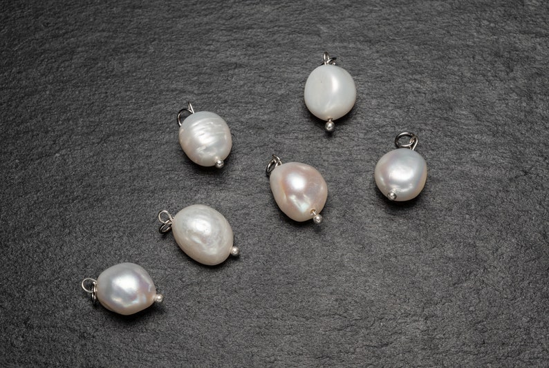 May include: Six white pearl charms with silver loops, displayed on a dark gray surface.