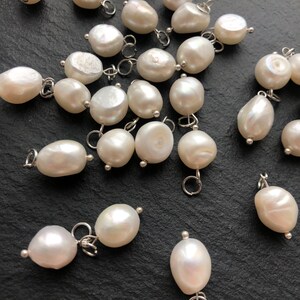 May include: A collection of white pearl charms with silver jump rings. The pearls are various shapes and sizes, and some have a slight iridescent sheen.