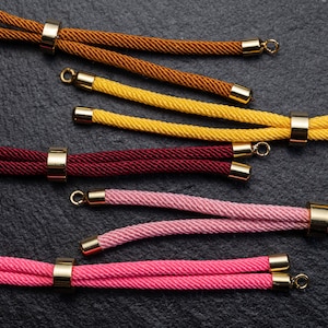 Op de afbeelding: Vijf verschillende gekleurde koorden met gouden metalen eindkappen en ringen. De koorden zijn bruin, geel, bordeauxrood, roze en lichtroze.