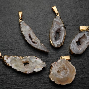 Könnte beinhalten: Fünf natürliche Achat-Anhänger mit goldfarbenen Ösen. Die Anhänger haben verschiedene Formen und zeigen grau-weiße Bänderung mit kristallgefüllten Hohlräumen. Die Kanten sind mit Gold verziert, was einen auffälligen Kontrast erzeugt.