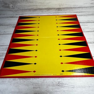 May include: A rectangular backgammon board featuring a bright yellow background with alternating black and red triangular points arranged in two rows. The board includes small printed text at the bottom center that reads 1972 Milton Bradley Co. 4319-X.