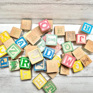 May include: A collection of wooden alphabet blocks featuring various letters in red, blue, green, and yellow paint. The square blocks are scattered in a pile, displaying different characters including A, B, C, D, F, H, I, J, L, M, N, O, P, Q, R, T, W, X, Y, and Z on their faces.