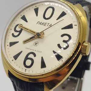 Peut inclure: Une montre-bracelet dorée avec un cadran crème présentant de grands chiffres noirs, des marqueurs triangulaires noirs et des aiguilles dorées. Le cadran affiche le texte PAKETA, un petit logo en étoile et le texte cyrillique СДЕЛАНО В СССР. Bracelet noir.