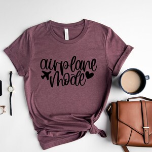 May include: A mauve colored t-shirt with the text "airplane mode" in black lettering. The shirt is folded and tied in a knot. A small airplane icon and two hearts are also included in the design.