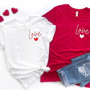 May include: Two white t-shirts with the word "Love" printed on them in red and white. The shirts are laid out on a white background with red hearts.