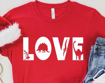 May include: Red t-shirt with white text that says "LOVE" with dinosaur silhouettes inside the letters. The shirt is laid out on a white surface with a Santa hat, a string of silver bells, and a pair of white sneakers.