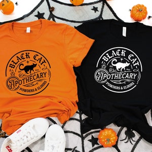 May include: Two t-shirts with a black cat and the text "Black Cat Apothecary Est. 1943 Powders & Elixirs" in a circle. The t-shirts are orange and black.