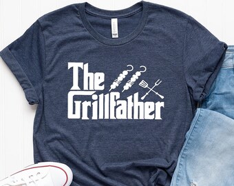May include: A navy blue t-shirt with white text that reads "The Grillfather" and a graphic of grilling tools. The shirt is laid out on a white surface with a pair of white sneakers and blue jeans.