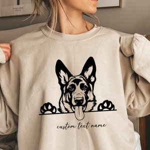 May include: A beige sweatshirt with a black silhouette of a German Shepherd dog peeking over the top. The dog has its tongue sticking out and paws visible. The text "custom text name" is printed below the dog.
