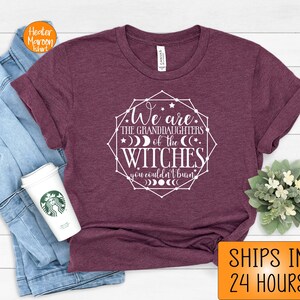 May include: Maroon t-shirt with the text "We are the granddaughters of the witches you couldn't burn" in a white geometric design. The shirt is folded, with a coffee cup and a denim jacket visible.