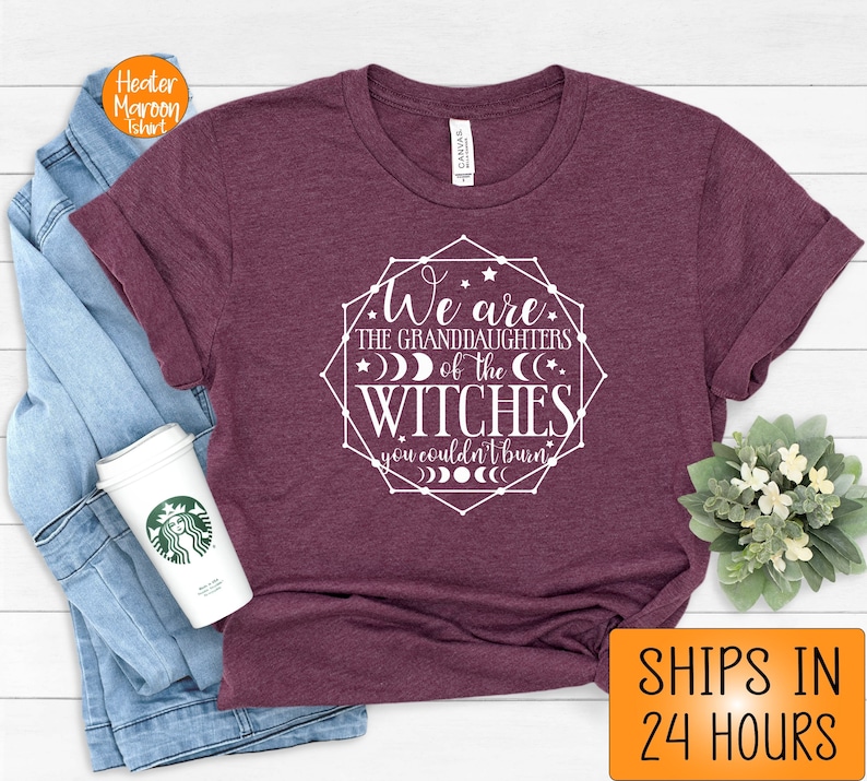 May include: Maroon t-shirt with the text "We are the granddaughters of the witches you couldn't burn" in a white geometric design. The shirt is folded, with a coffee cup and a denim jacket visible.