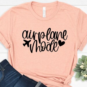 May include: A light pink t-shirt with the text "airplane mode" in black cursive lettering. A small airplane icon and a heart are included in the design.