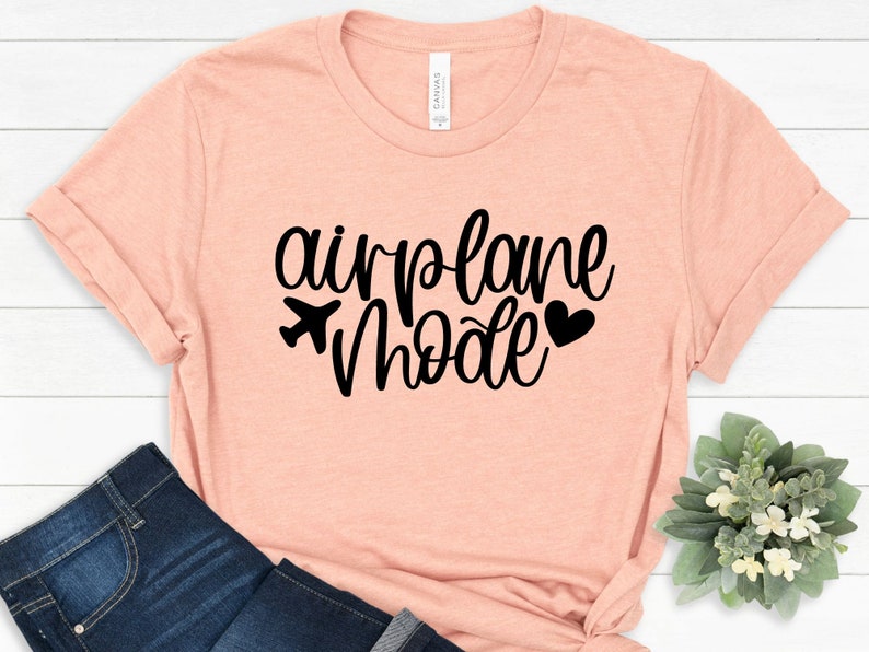 May include: A light pink t-shirt with the text "airplane mode" in black cursive lettering. A small airplane icon and a heart are included in the design.