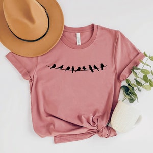 May include: A dusty rose t-shirt with a graphic of a line of black birds on a wire. The t-shirt is made of a soft, comfortable material.