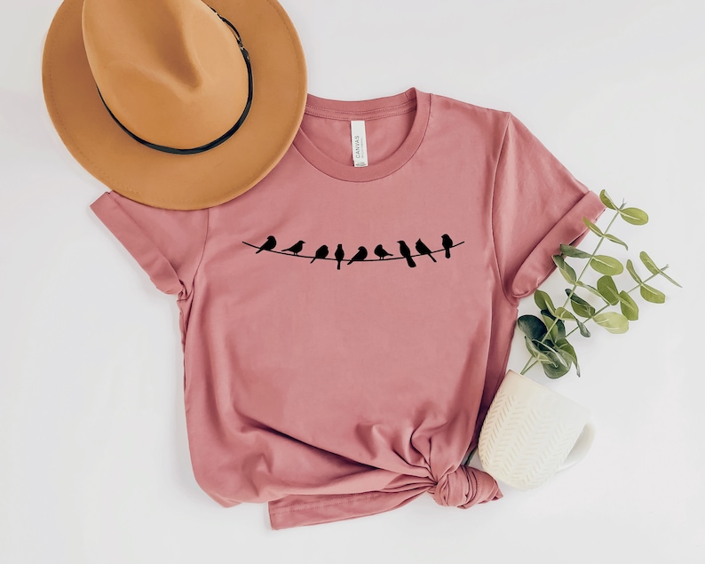 May include: A dusty rose t-shirt with a graphic of a line of black birds on a wire. The t-shirt is made of a soft, comfortable material.