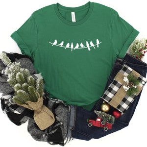 May include: A green t-shirt with a white silhouette of a line of birds on a wire. The shirt is folded and displayed with a plaid scarf, a pair of blue jeans, and a small red toy truck.