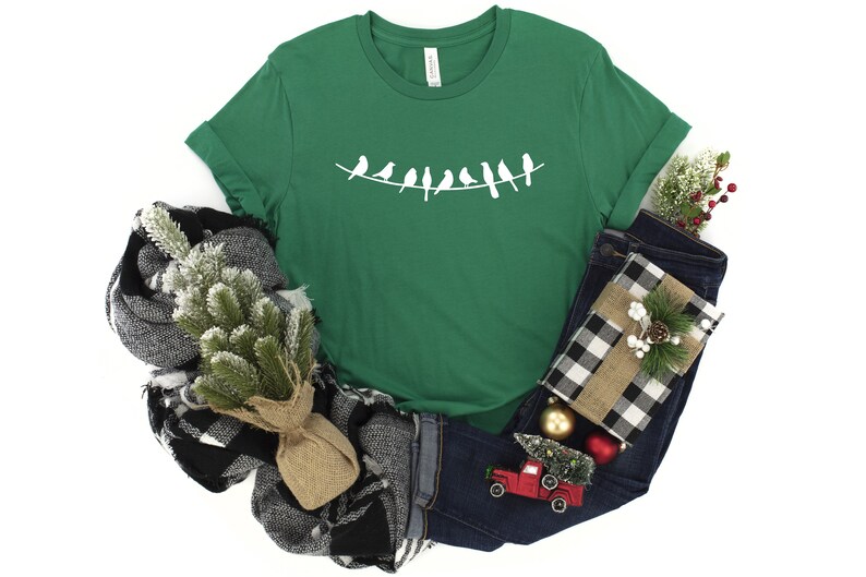 May include: A green t-shirt with a white silhouette of a line of birds on a wire. The shirt is folded and displayed with a plaid scarf, a pair of blue jeans, and a small red toy truck.
