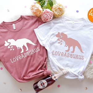 May include: Two t-shirts with a dinosaur graphic and the text "Loveasaurus". One shirt is white with a pink dinosaur and the other is a heather mauve color with a white dinosaur.